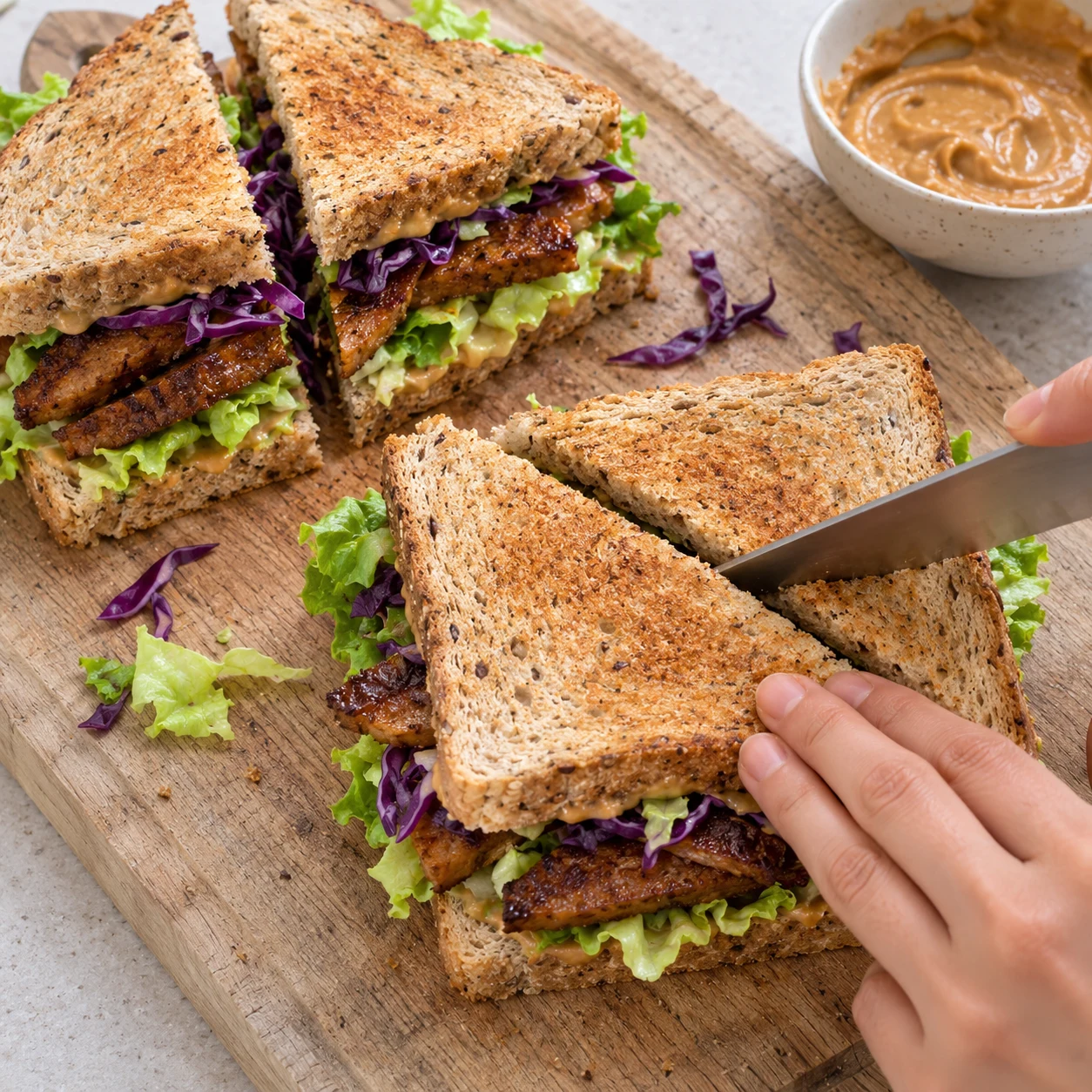 Tranche de pain de mie grillée tartinée de sauce cacahuète sur une planche en bois, garnie de salade verte et de lamelles de steak végétal dorées posées avec des baguettes, mains adultes anonymes.