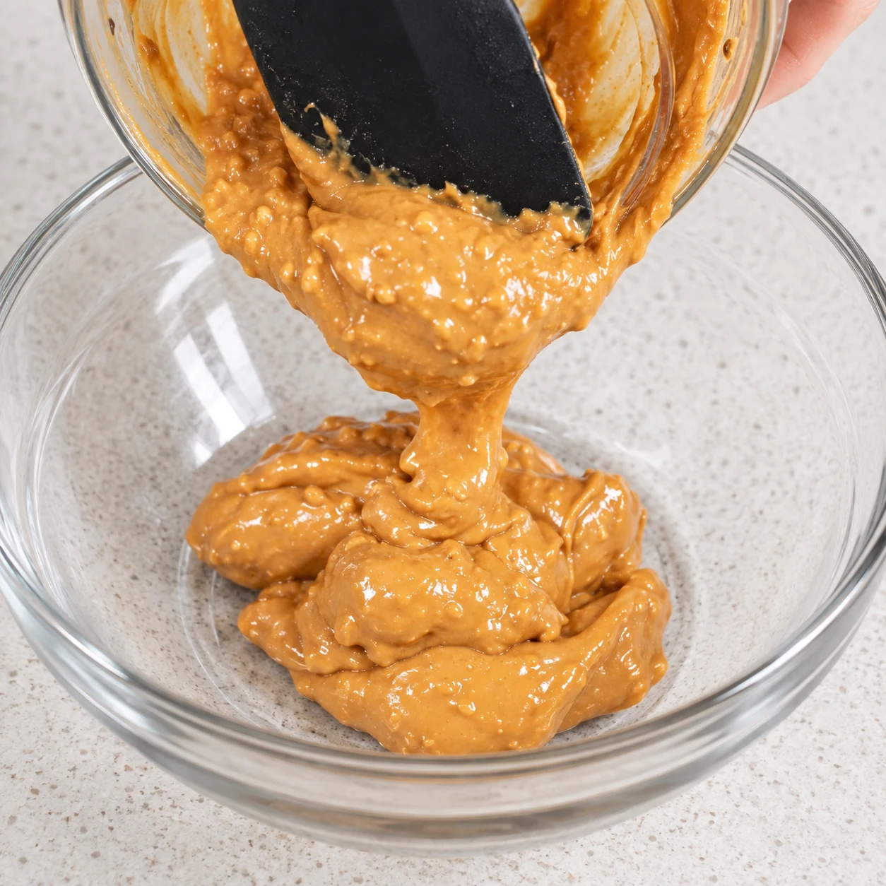 Spatule raclant la purée de cacahuètes épaisse, brun doré, dans un saladier en verre ; aucun flocon ni sirop encore ajouté.