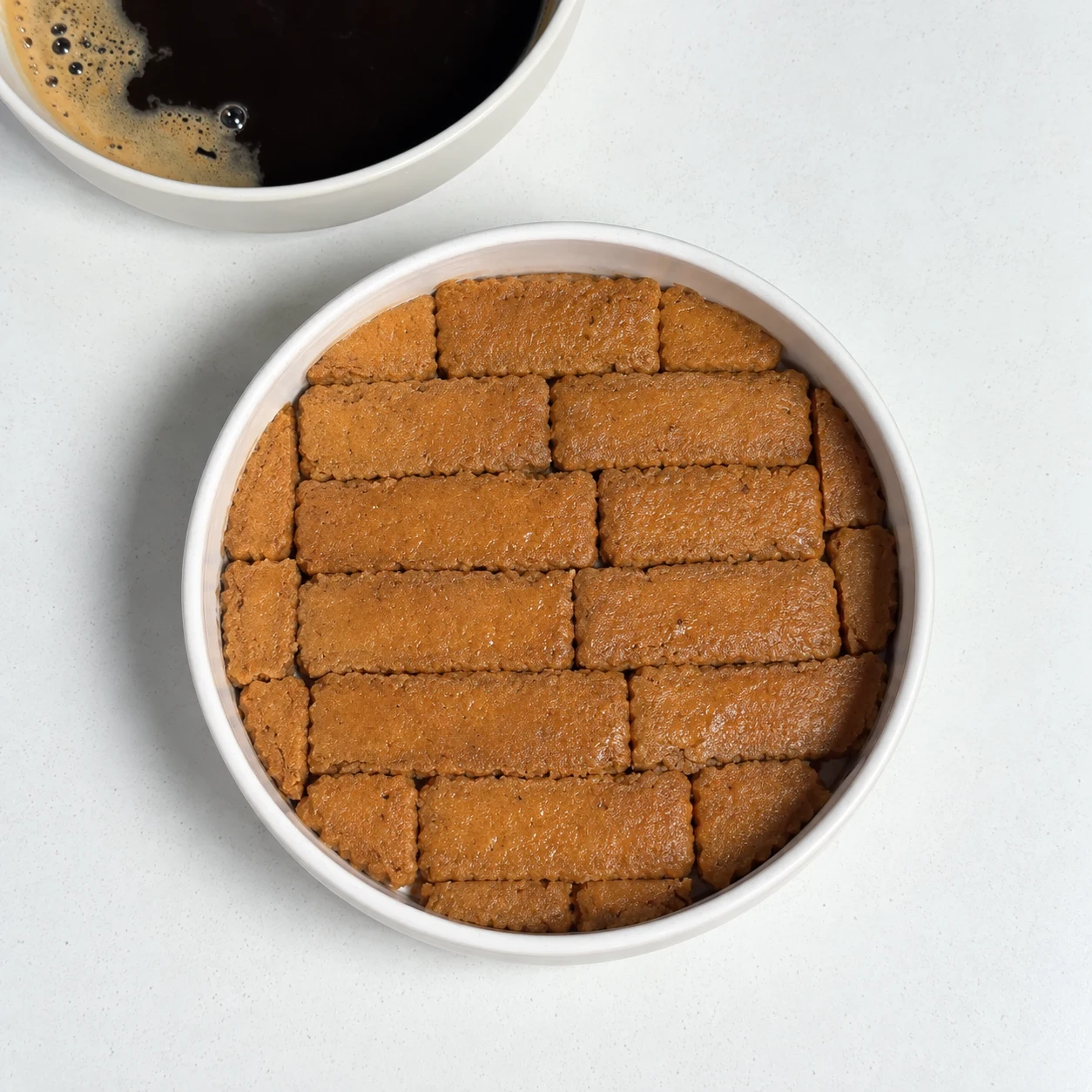 Plat rond en céramique blanche dont le fond est couvert de spéculoos imbibés de café, rangés côte à côte.