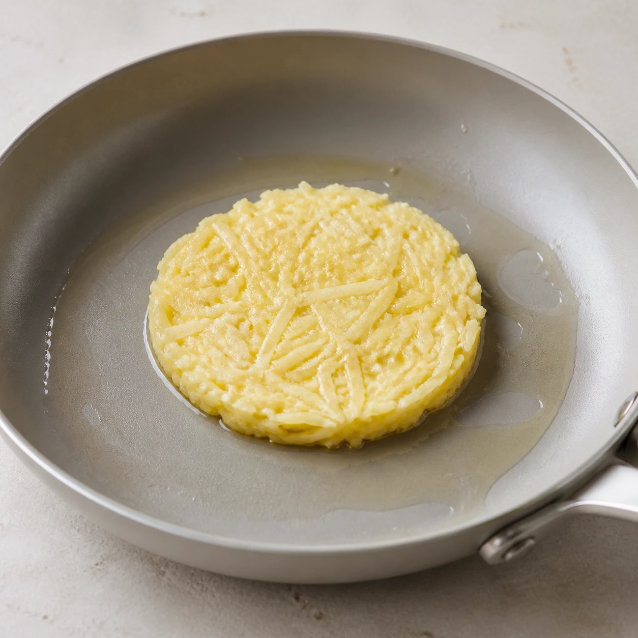 Une petite galette compacte de pomme de terre crue est tassée dans une poêle chaude légèrement huilée.