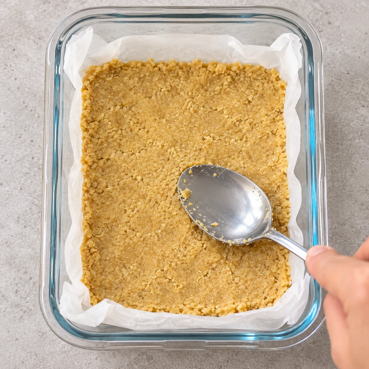 Une cuillère presse la pâte granuleuse claire au fond d’un petit plat rectangulaire chemisé.