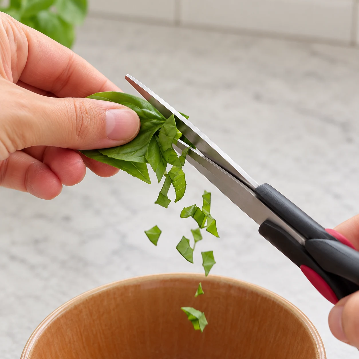 Gros plan carré sur des mains adultes anonymes qui cisaillent les feuilles de basilic frais au-dessus du bol de sauce, comme à 26 s. Cadrer les ciseaux et le basilic : le contenu du bol reste entièrement hors champ. Aucun autre ingrédient visible.