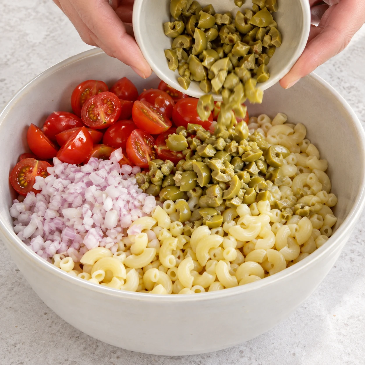 Saladier de coquillettes cuits et refroidis avec demi-tomates cerises, échalote et olives hachées avant sauce.