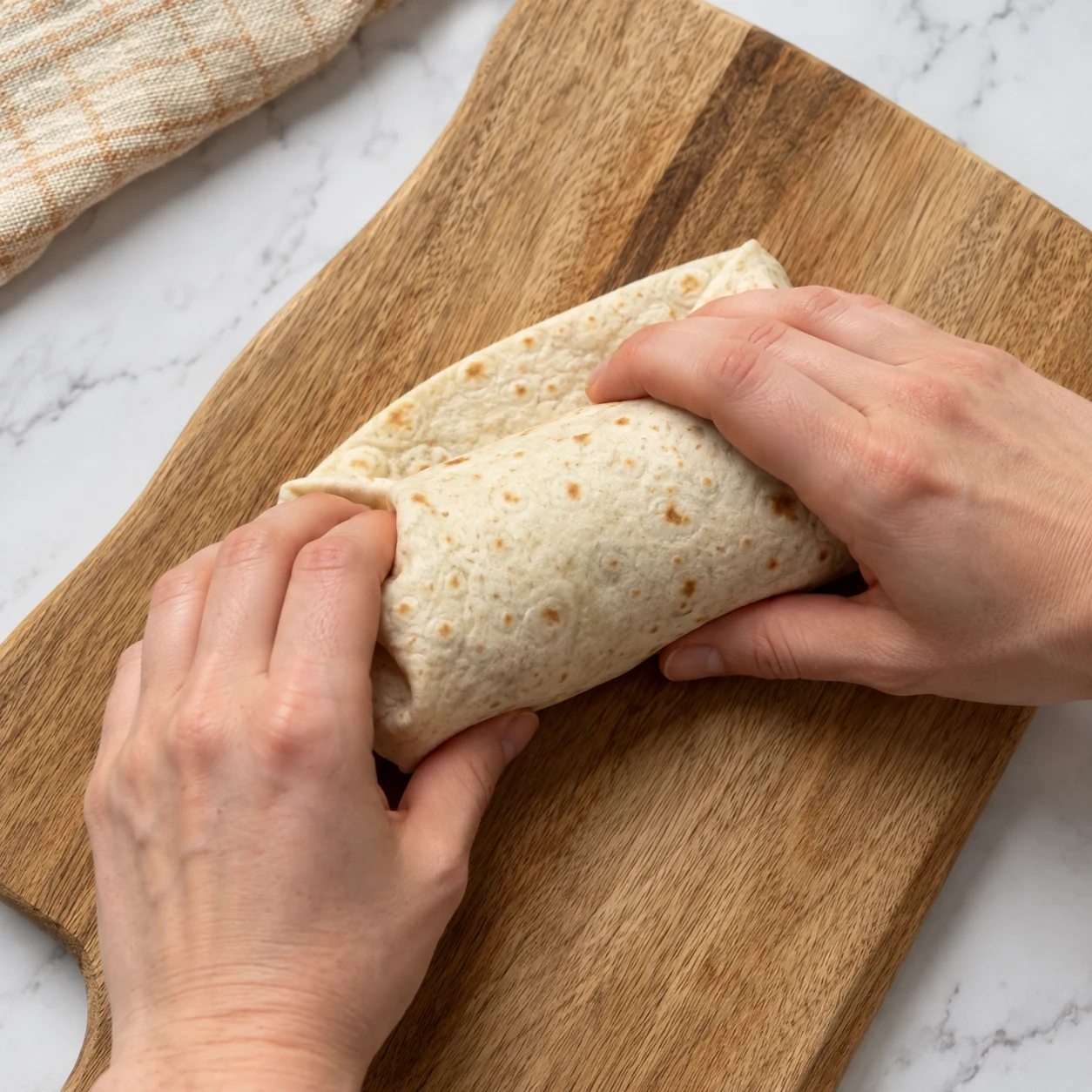 Deux mains adultes anonymes rabattent et roulent une tortilla garnie, côtés visibles et rentrés.