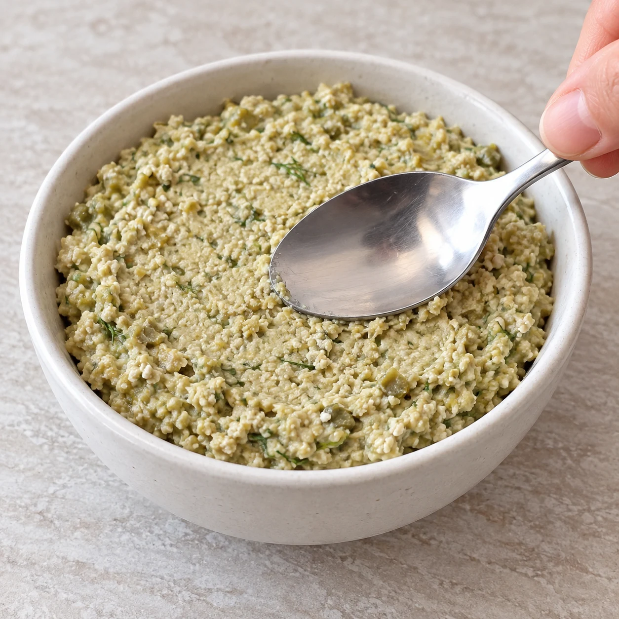 Dos d’une cuillère égalisant une tapenade granuleuse vert pâle dans un petit bol neutre.