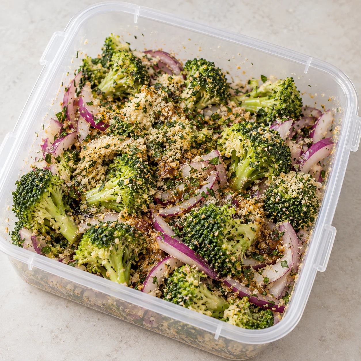 Boîte transparente contenant brocoli cru, oignon rouge et enrobage sec légèrement huilé.