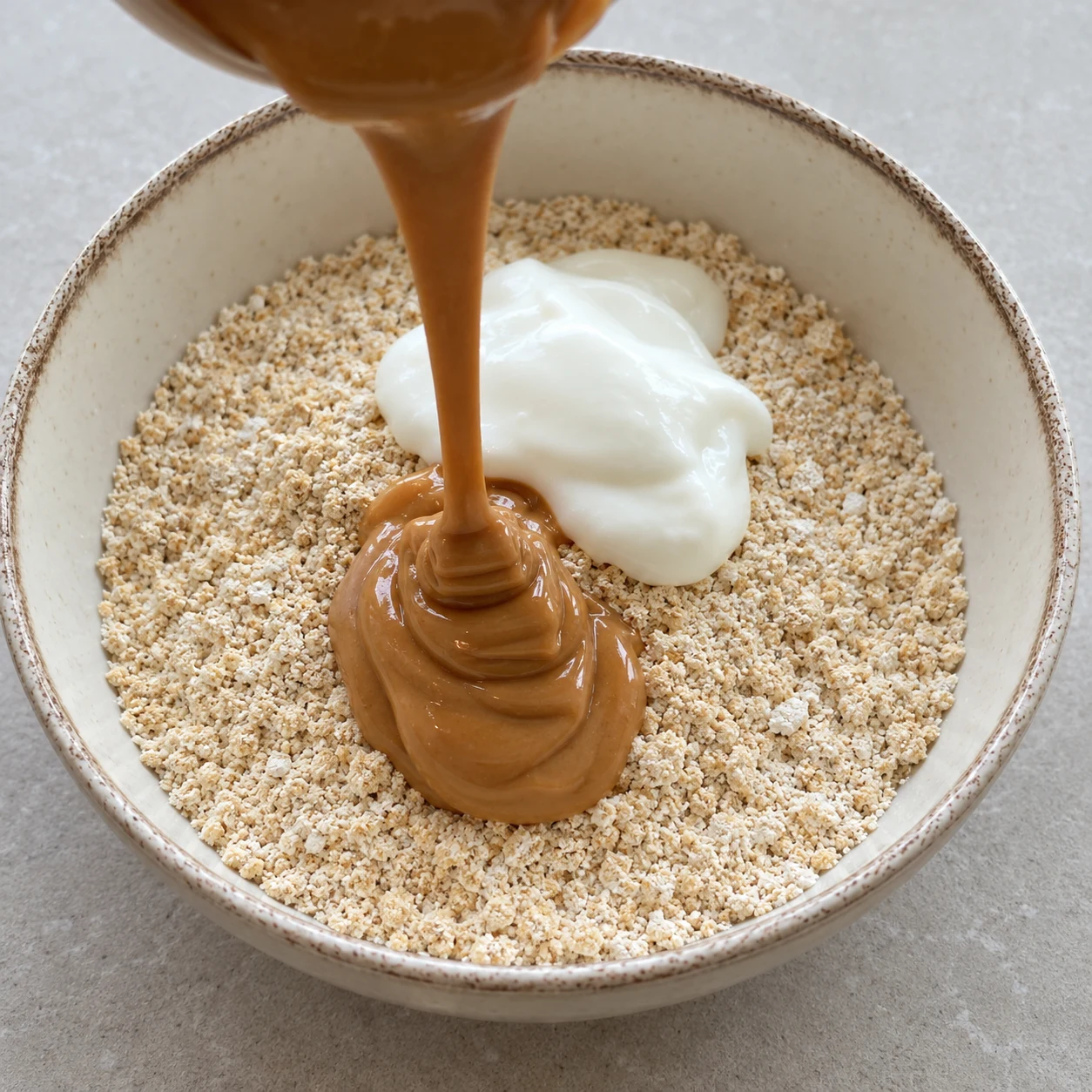 Purée de cacahuète brune versée en filet épais sur une poudre beige d'avoine et une cuillerée de yaourt de soja blanc, dans un saladier en céramique.