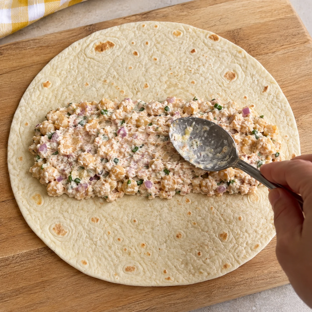 Une cuillère étale la garniture crémeuse aux pois chiches en bande centrale sur une tortilla de blé ouverte. Aucun légume de garniture à cette étape.
