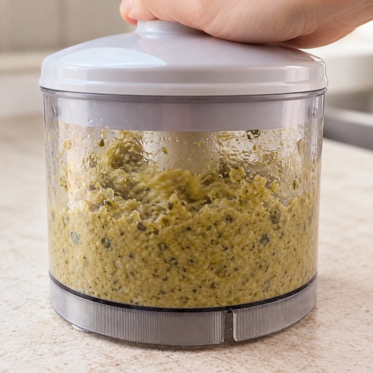 Petit mixeur transparent fermé en fonctionnement, tapenade jaune-vert humide en cours de broyage, légèrement granuleuse, une main adulte anonyme presse le couvercle. Aucun morceau entier de fruit ni pain.