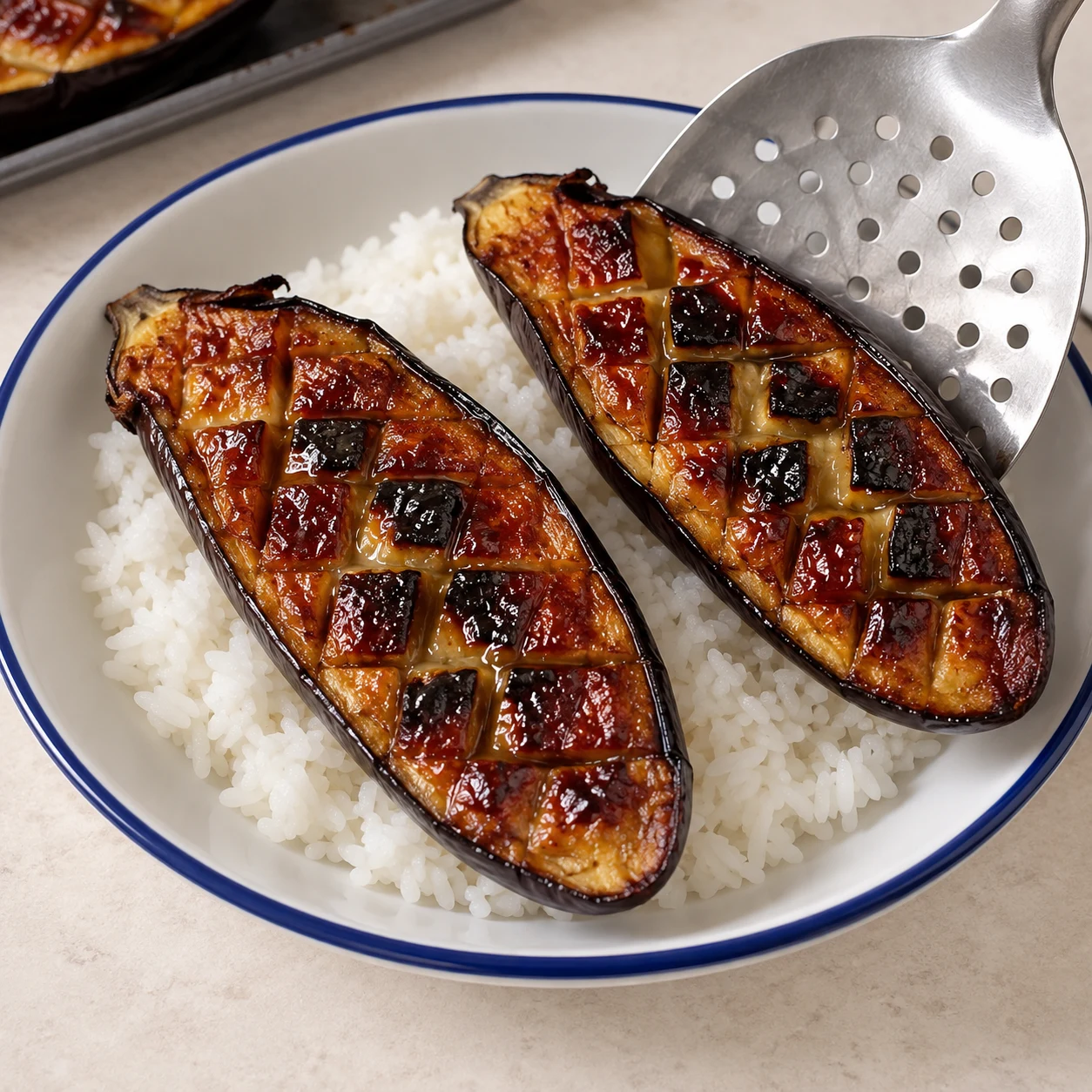 Spatule déposant une demi-aubergine rôtie laquée sur du riz ; aucun topping encore.