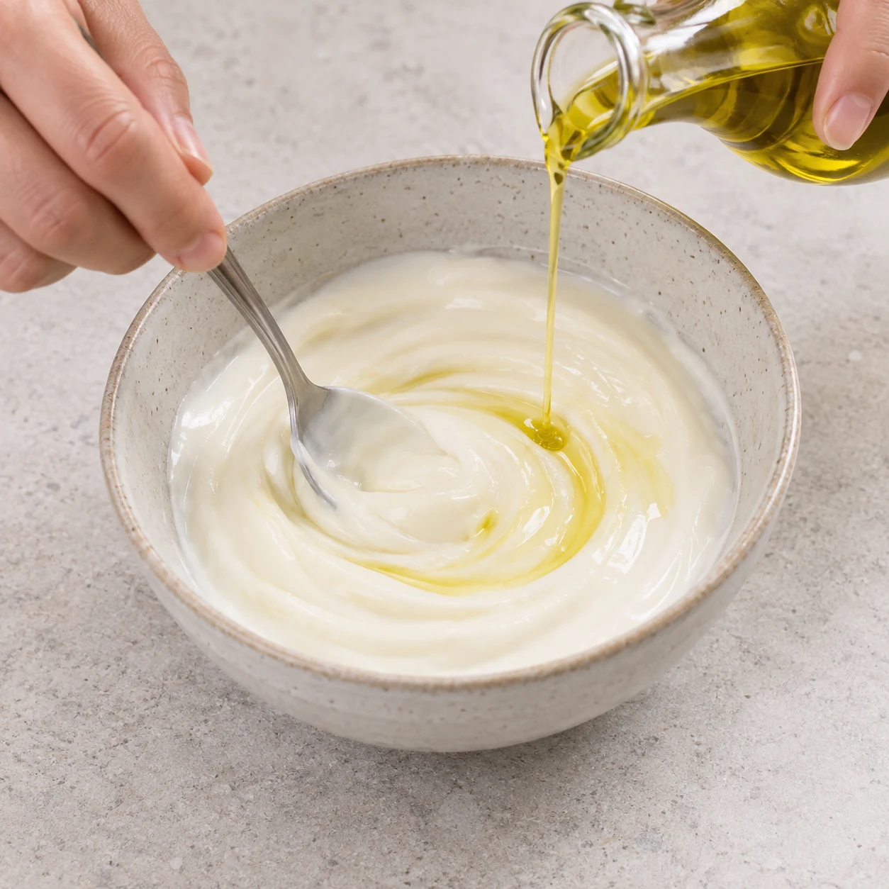 Yaourt de soja blanc crémeux dans un bol neutre, main mélangeant avec une cuillère et fin filet d’huile.