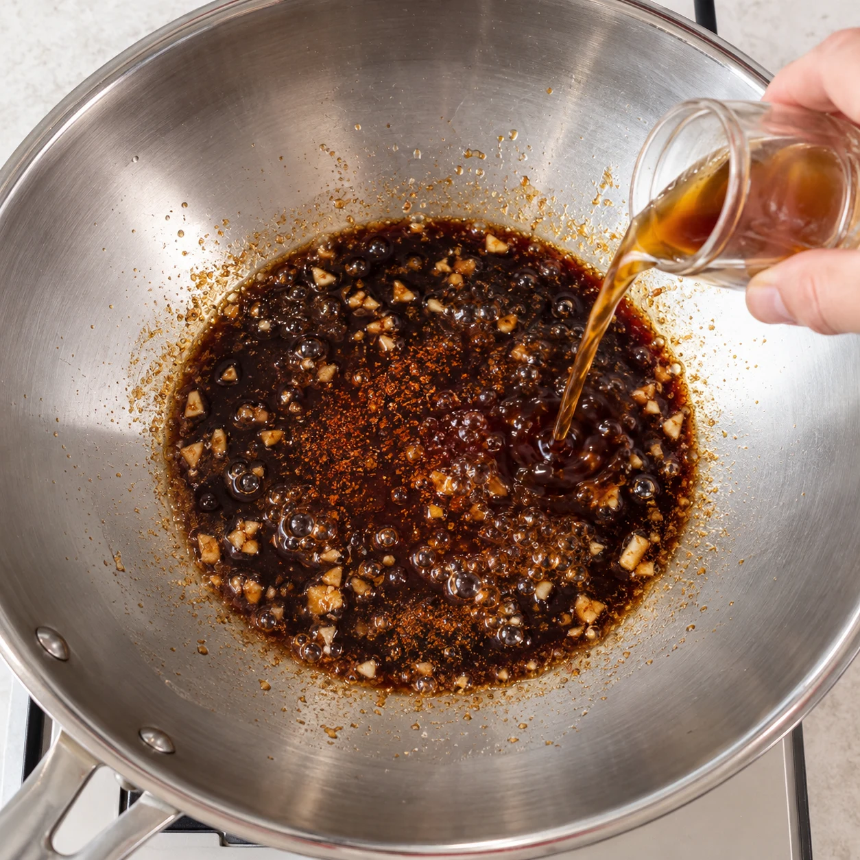 Une cuillère ajoute un condiment liquide dans la base sombre ail et sauce soja réduite en sel, wok en inox.