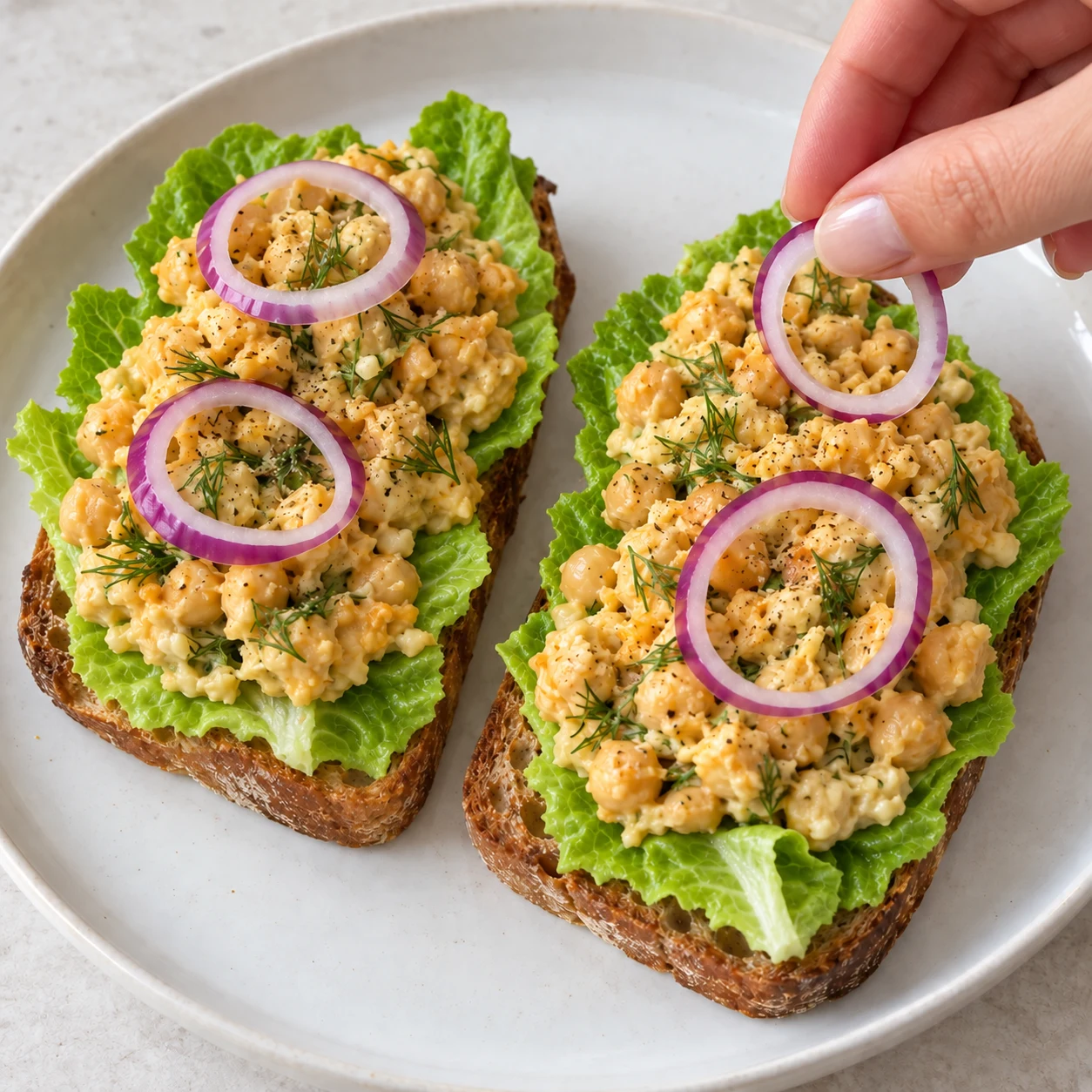 Main adulte anonyme déposant des rondelles d’oignon rouge sur un toast au pois chiches crémeux à la sriracha et à l’aneth.