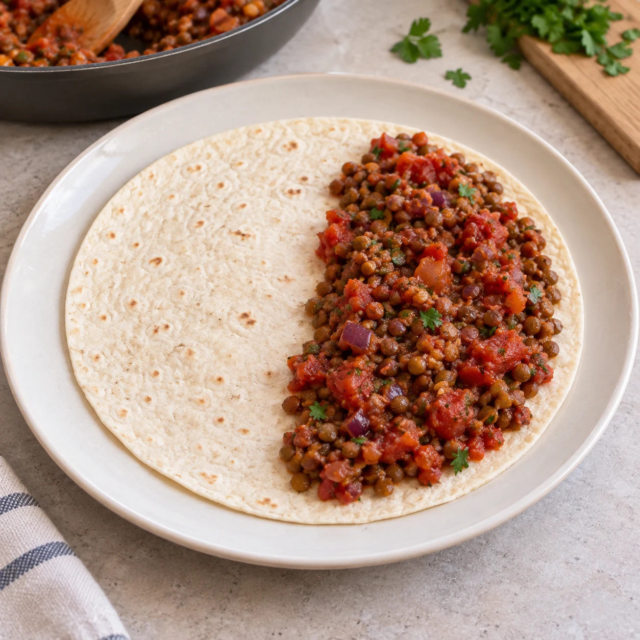 Photographie pédagogique carrée : Pose la tortilla dans une assiette et dépose une part de garniture sur une moitié. Répartis la garniture entre les six tortillas. Résultat : La garniture occupe une moitié et laisse l’autre libre pour le pliage. Mains adultes anonymes seulement si nécessaires ; lentilles cuites petites et brunes à la place des haricots rouges de la capture, option source documentée ; fromage végétal au lieu de laitier lorsque présent. Aucun texte.