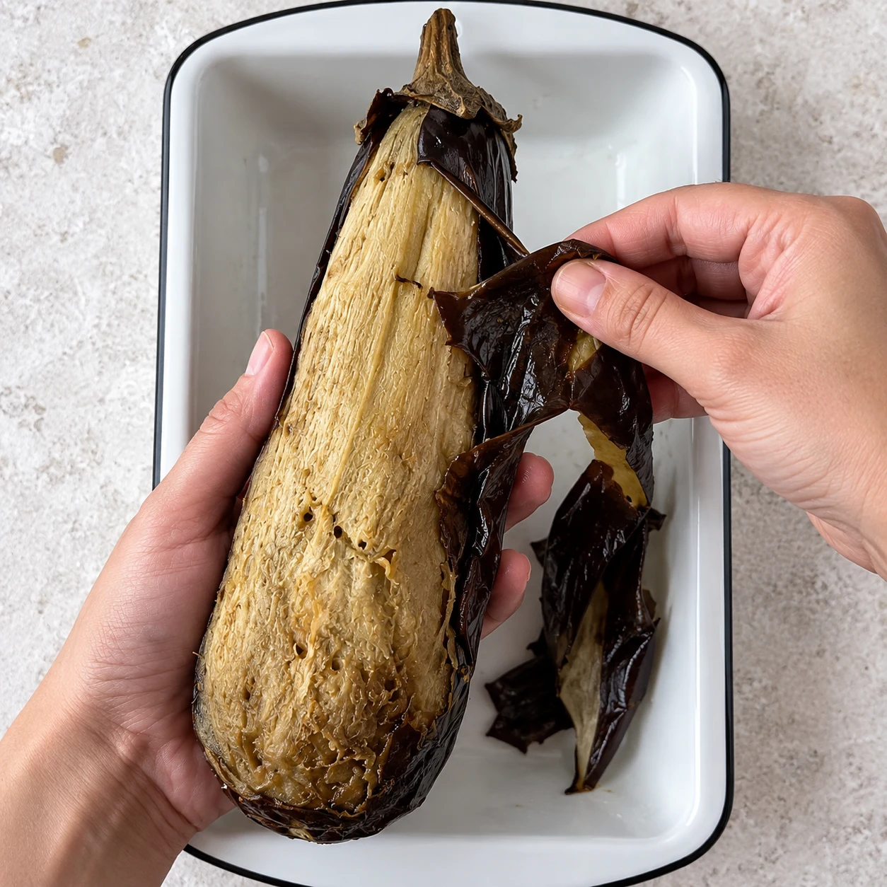 Laissez l’aubergine refroidir suffisamment pour la manipuler, puis retirez sa peau avec les mains en conservant le pédoncule. Montrer une seule phase de préparation, sans texte et sans personne identifiable.