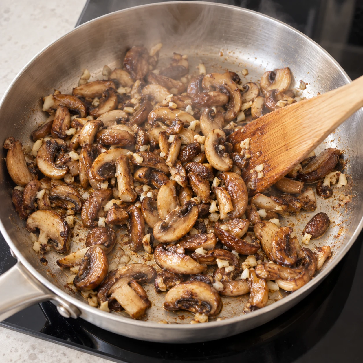 Champignons bruns et ail dans la poêle inox, vapeur et dernières gouttes d’eau, spatule en bois.