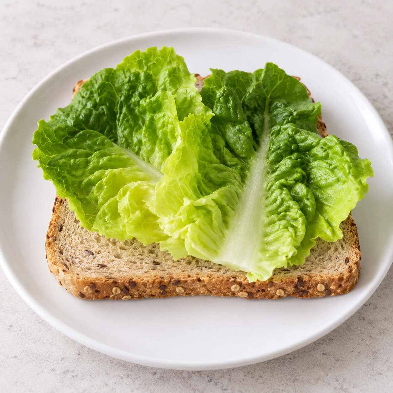 Déposez 2 à 3 feuilles de salade verte propres et sèches sur une tranche de pain de mie. Montrer uniquement les ingrédients de cette phase sur pierre pâle, vaisselle blanche neutre.