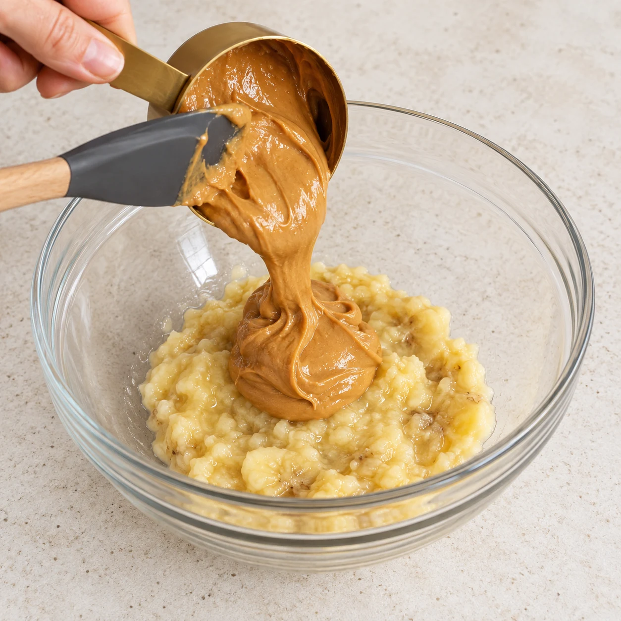 Déposez les 60 g de beurre de cacahuète sur la purée de banane. Une seule phase, texture fidèle à la recette d’origine, plan de travail pierre pâle et vaisselle neutre ; mains anonymes seulement.