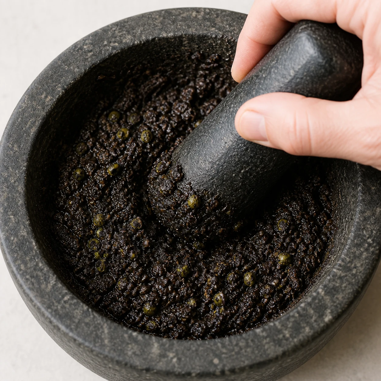 Câpres incorporées à une pâte d’olives noire dans un mortier, texture fine mais légèrement granuleuse.