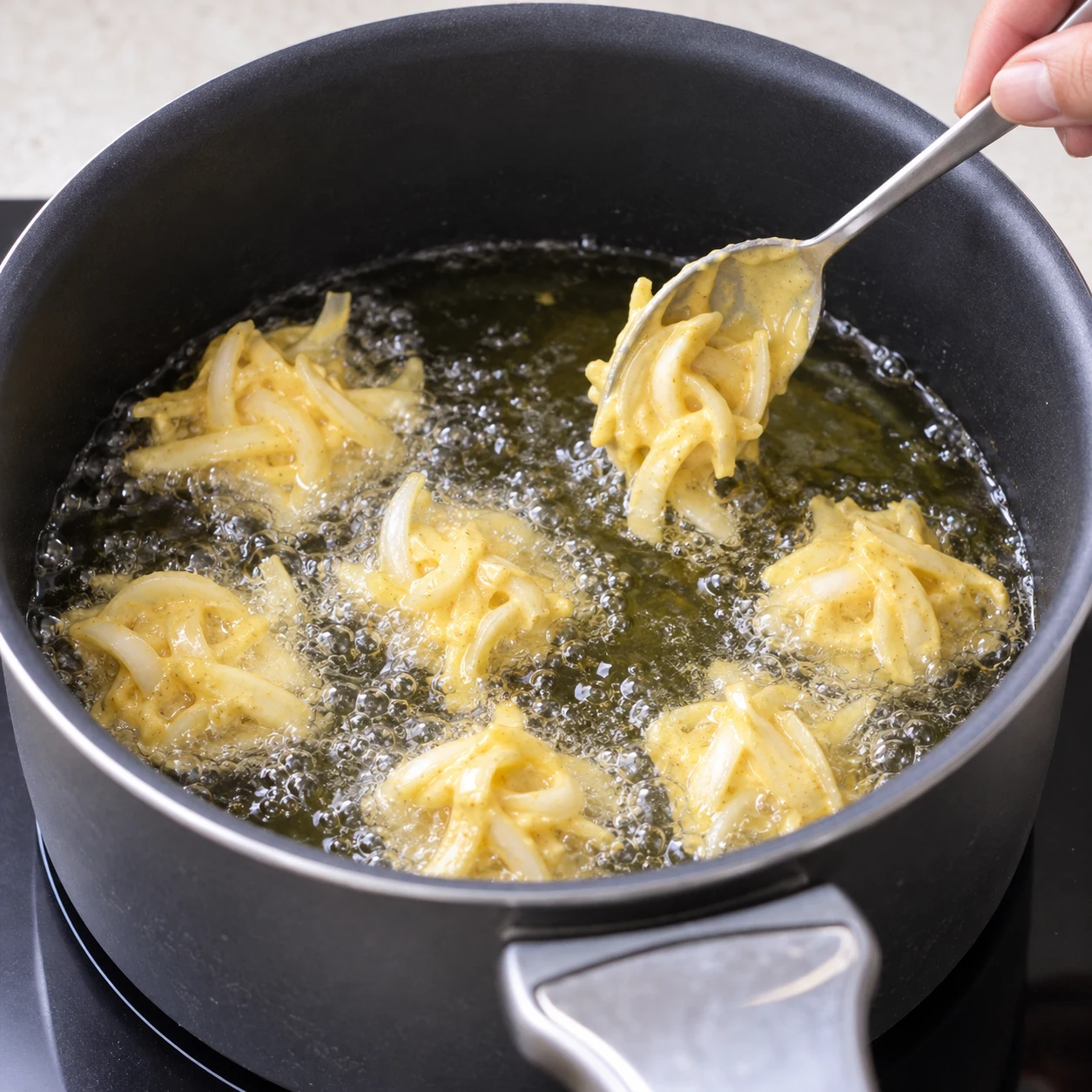 Une cuillère dépose un petit amas d’oignon enrobé de pâte jaune pâle au ras d’un bain d’huile frémissant dans une casserole noire ; début de friture, aucun beignet déjà brun.