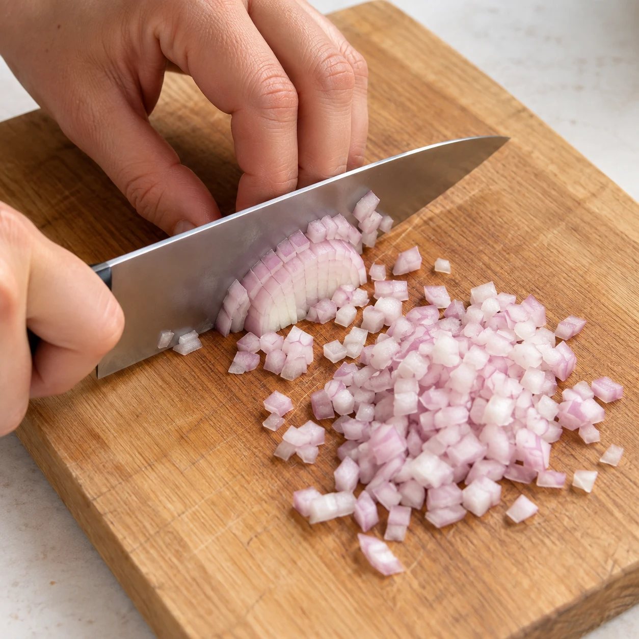 Couteau et main en griffe ciselant une échalote blanche légèrement violette en petits dés sur une planche, aucun autre aliment.