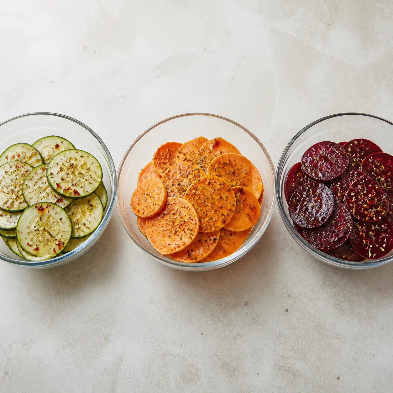 Trois bols en verre alignés contenant séparément des rondelles de courgette, des tranches de patate douce et des tranches de betterave, chaque bol brillant d'huile d'olive avec sel, poivre, piment d'Espelette rouge et herbes de Provence séchées visibles en surface, aucun autre ingrédient, aucun texte, aucune marque.