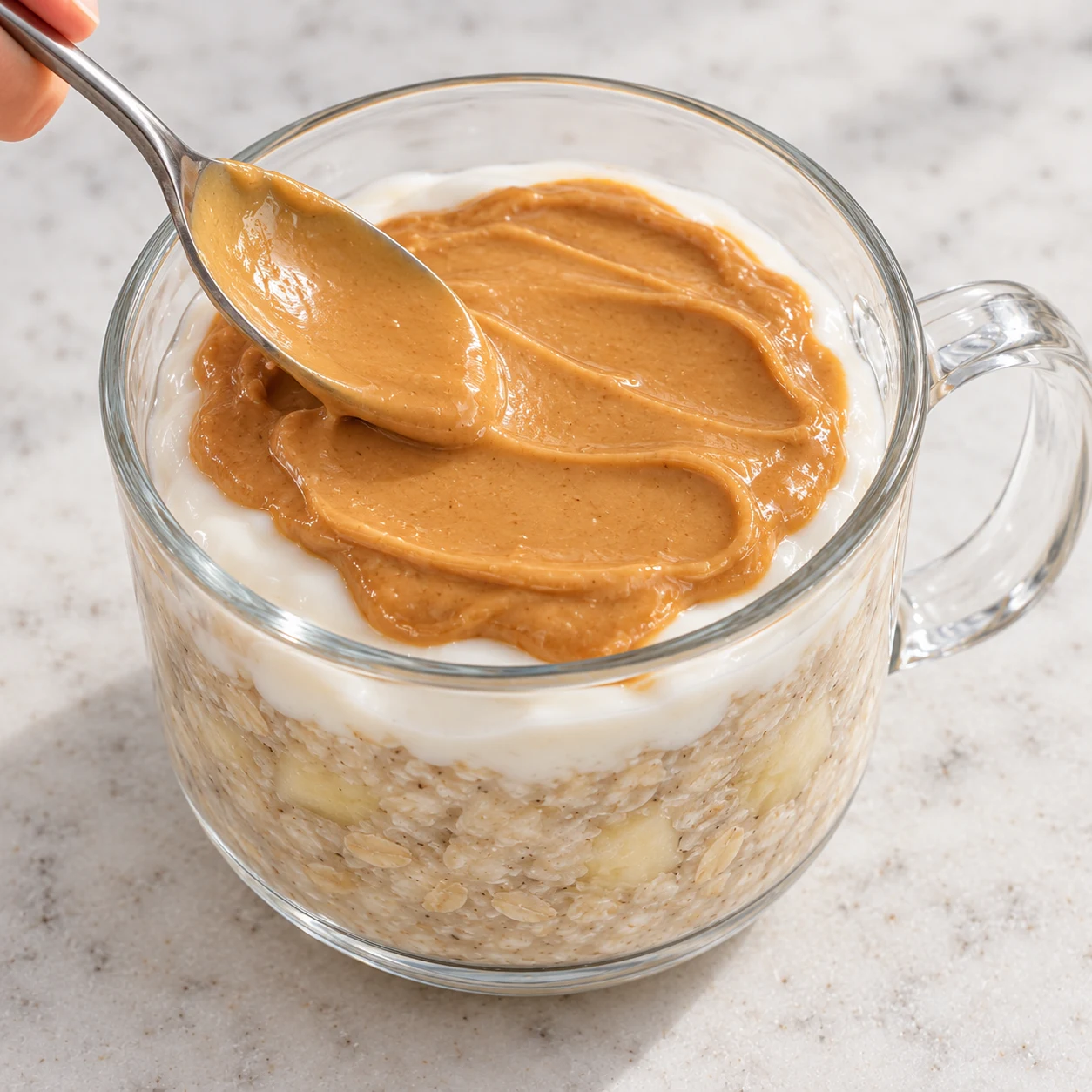 Étalez la cuillère à soupe de purée de cacahuète sur le yaourt, comme dans la recette d’origine. Montrer seulement cette phase de la variante à l’avoine, sans graines ni poudre, un récipient transparent à poignée sur pierre pâle, sans texte ni personne identifiable.