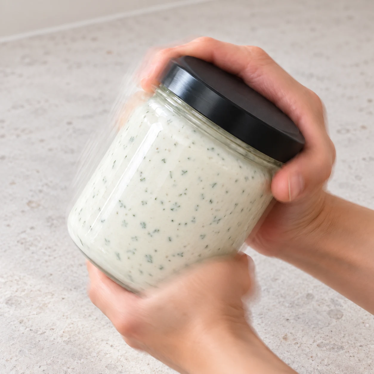 Mains adultes anonymes tenant et secouant un bocal fermé, sauce blanche aux herbes.
