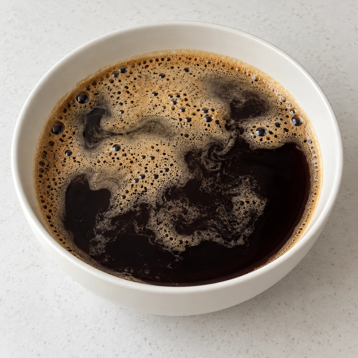 Bol large en céramique blanche rempli de café noir froid, légère mousse brune en surface, sur un plan de travail clair.