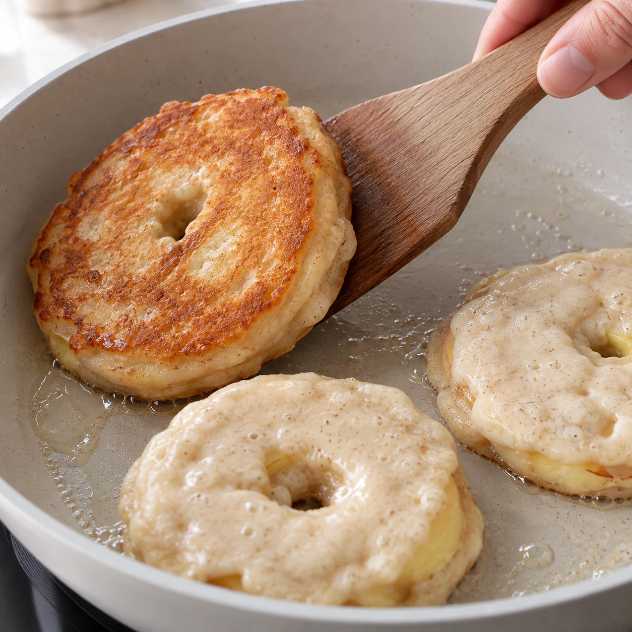 Spatule en bois retournant un beignet de pomme dans la poêle, une face brun doré et une face pâle.