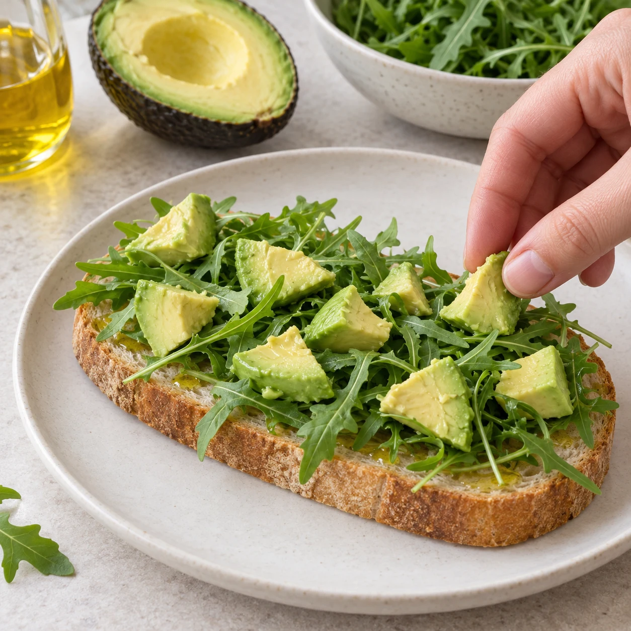 Répartis les morceaux du demi-avocat sur la roquette. Photographie carrée de cuisine, ingrédients végétaux finaux seulement, mains adultes anonymes si nécessaires, aucun texte ni visage.