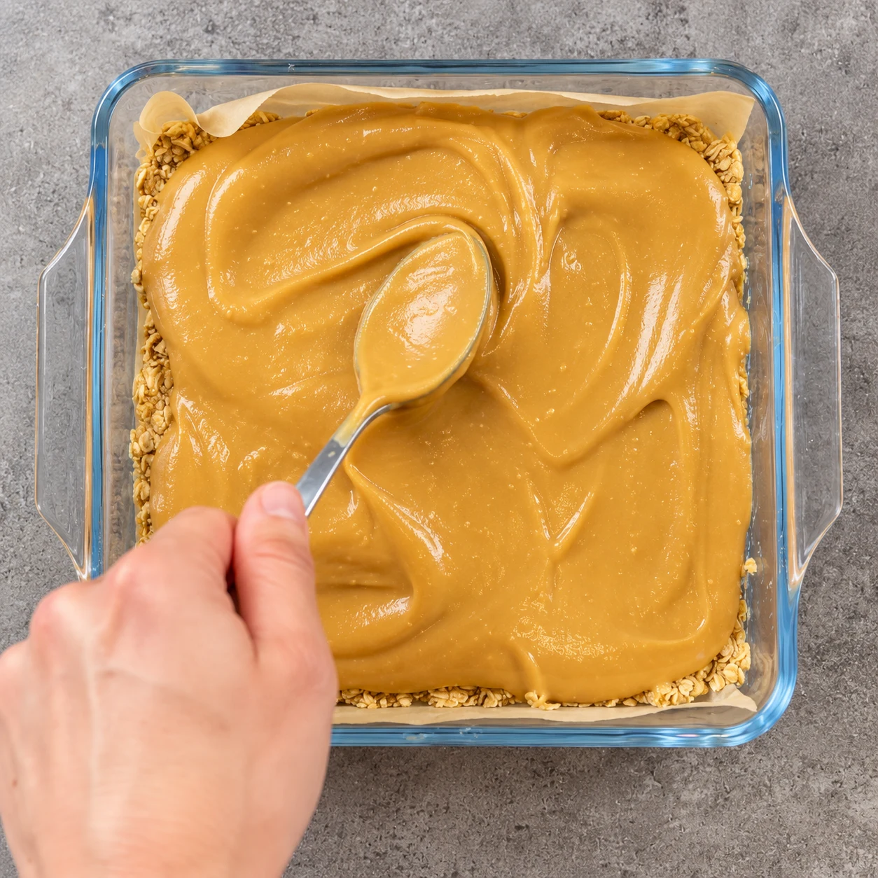 Cuillère étalant une couche épaisse de caramel cacahuète doré sur le croquant, dans le même moule carré chemisé.