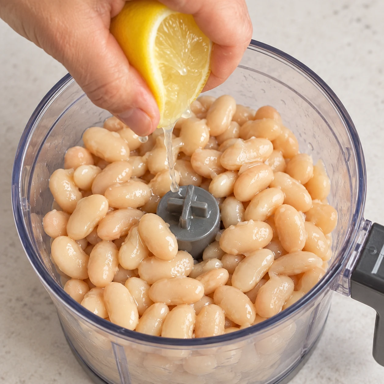 Main adulte anonyme pressant un demi-citron jaune au-dessus de haricots blancs cuits dans un bol de mixeur débranché, pas de peau dans le bol.
