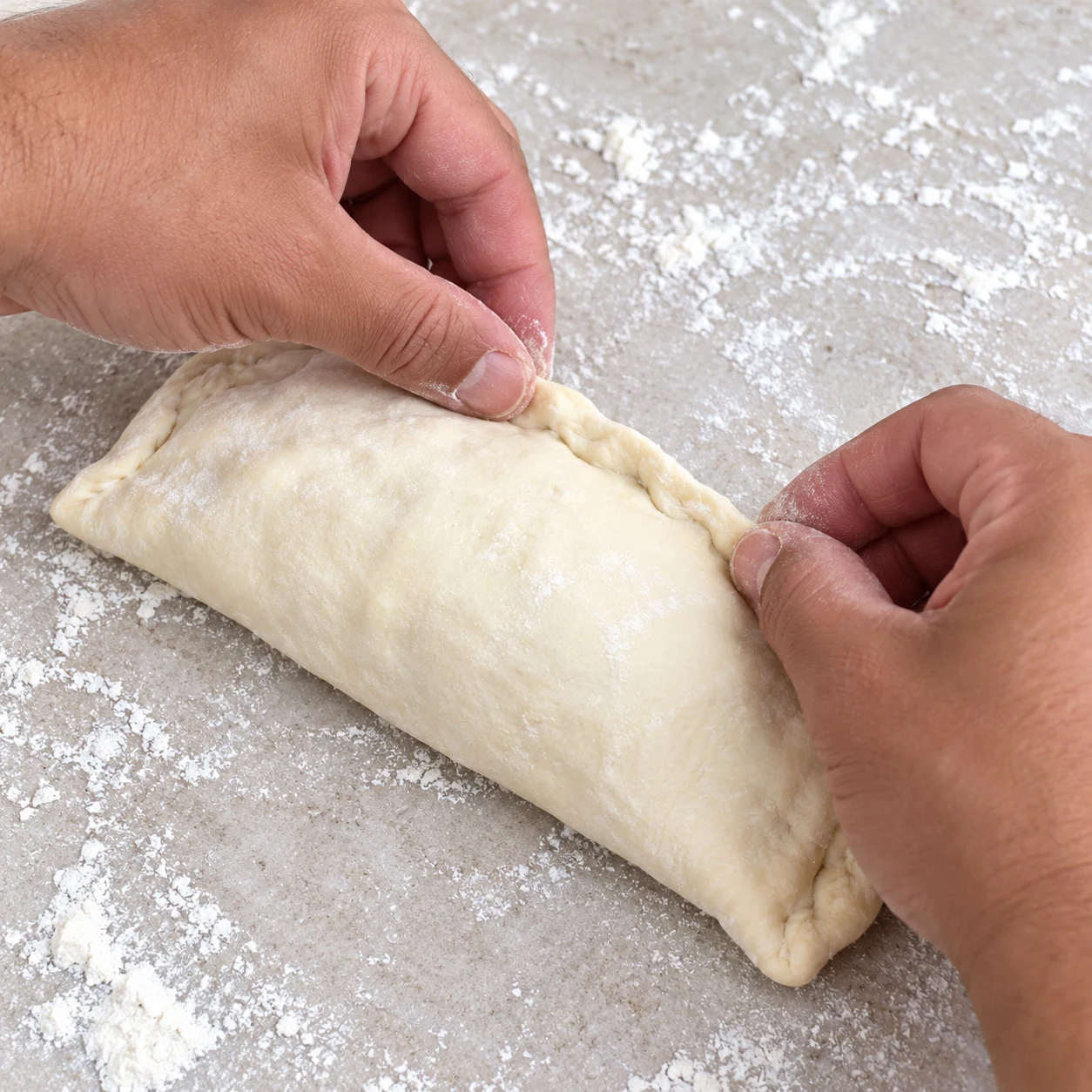 Deux mains anonymes pincent la soudure d’un panini cru sur pierre pâle.