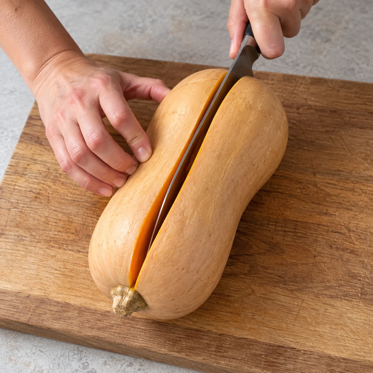 Coupe longitudinale d’une butternut crue sur planche stable, mains anonymes, tenue sûre, aucun autre aliment.