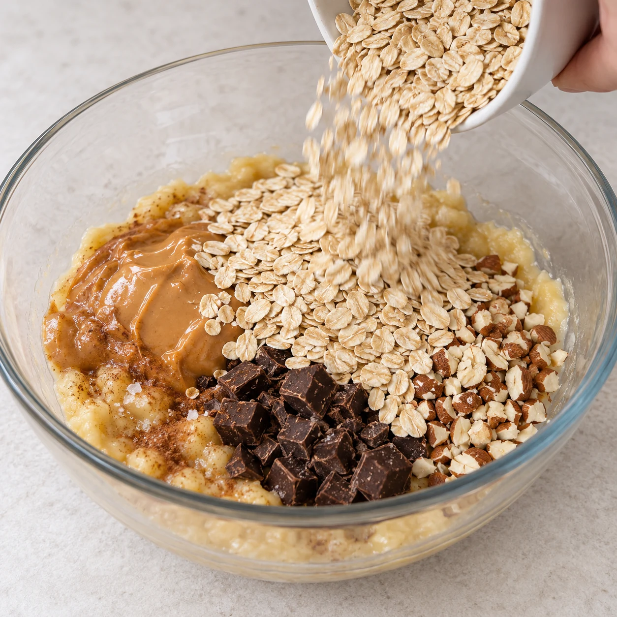 Ajout de flocons d’avoine au saladier contenant banane et beurre de cacahuète, chocolat et amandes, sel et cannelle discrets.