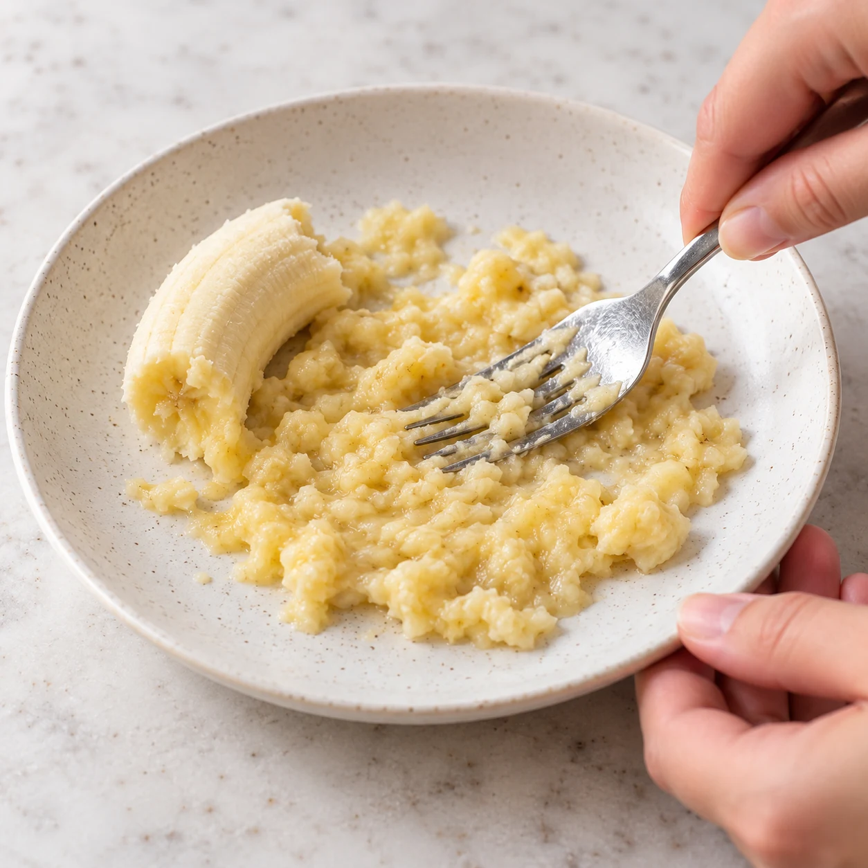 Écrasez la demi-banane à la fourchette dans une assiette. Montrer seulement cette phase de la variante à l’avoine, sans graines ni poudre, un récipient transparent à poignée sur pierre pâle, sans texte ni personne identifiable.