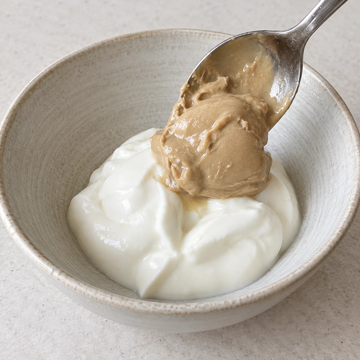 Cuillère déposant un tahini beige épais sur yaourt de soja nature blanc onctueux dans un bol neutre.