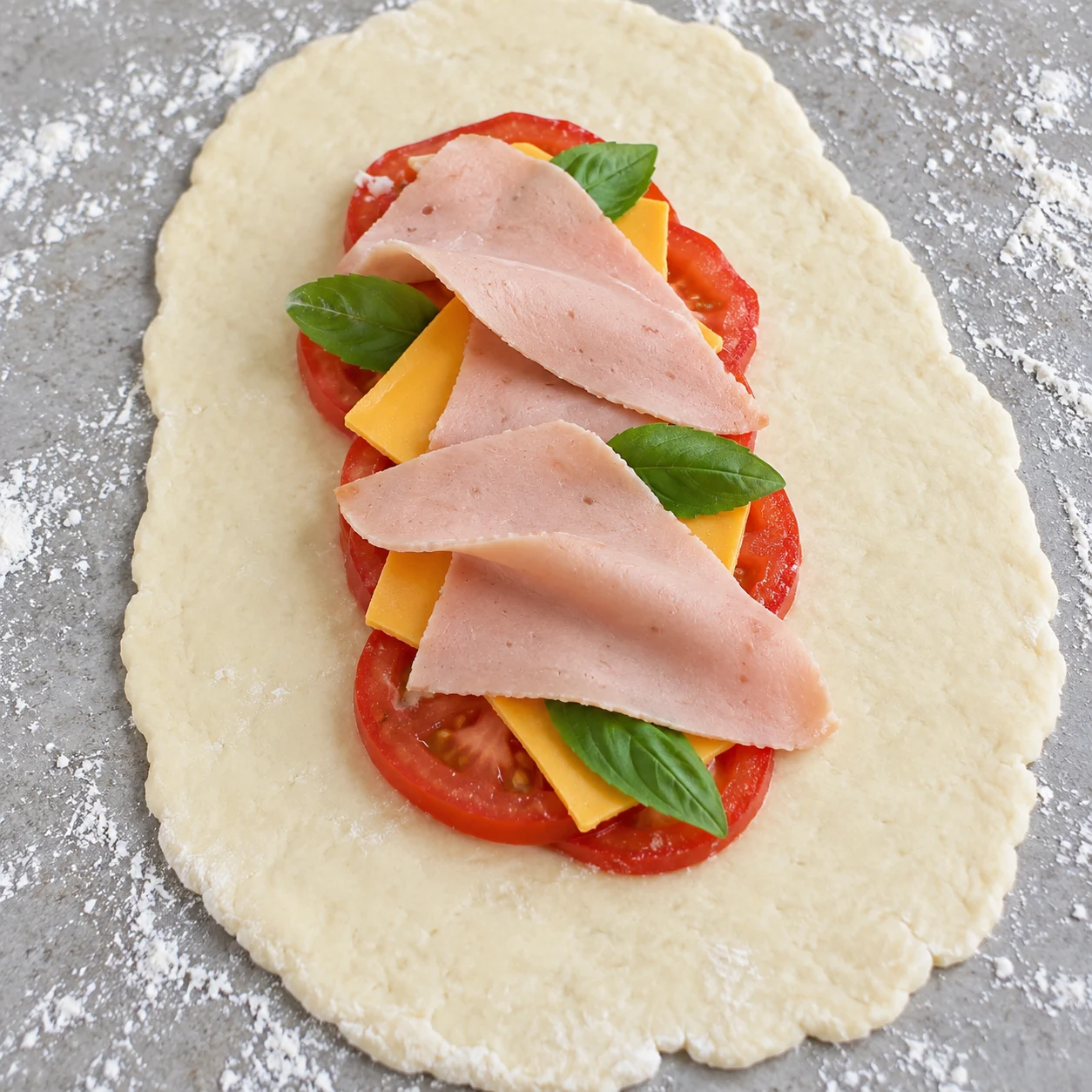 Une seule abaisse allongée garnie de tomate rouge, fines tranches de fromage végétal, basilic frais puis jambon végétal, mains anonymes.