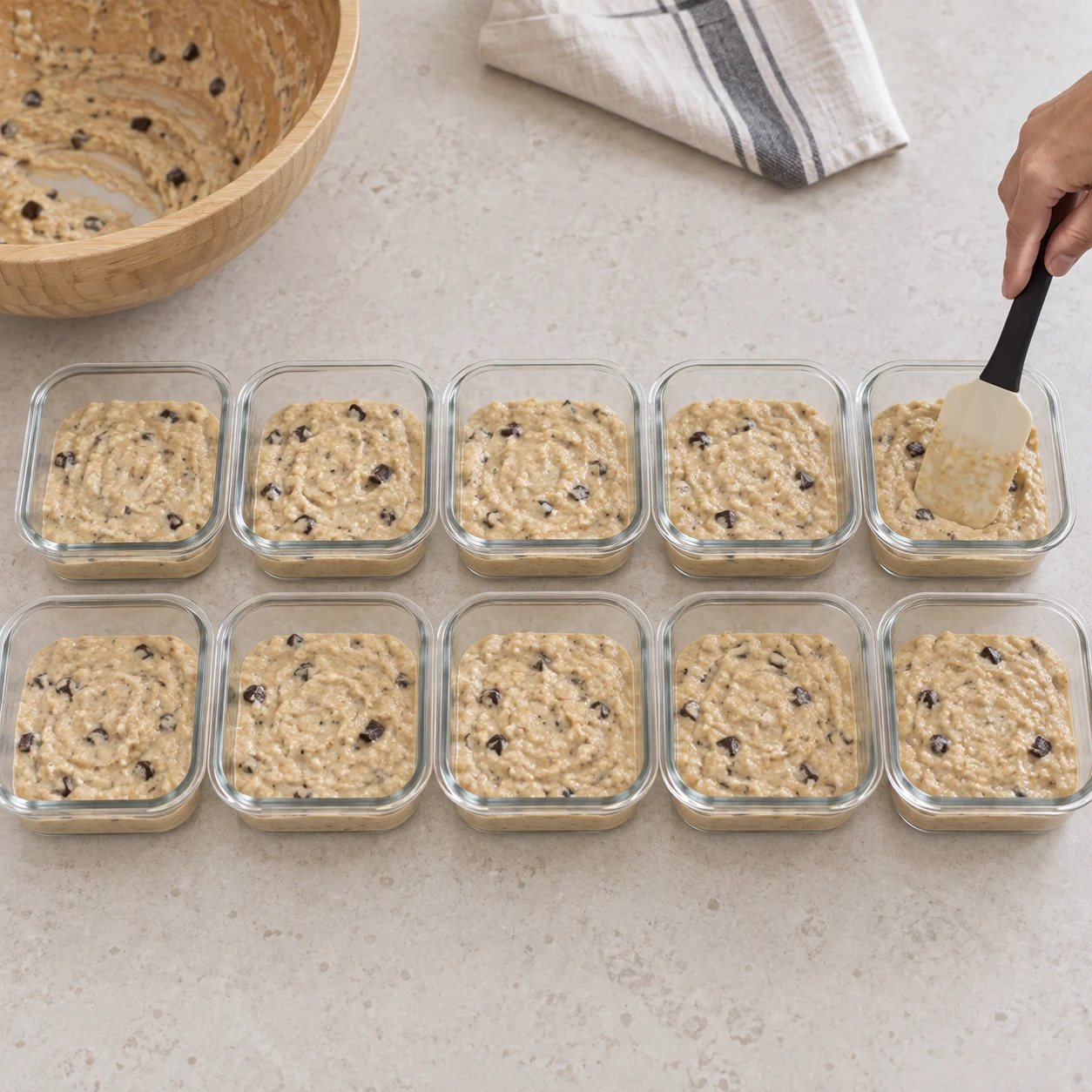 Une spatule répartit une pâte beige aux morceaux de chocolat dans des petits plats carrés en verre compatibles four.