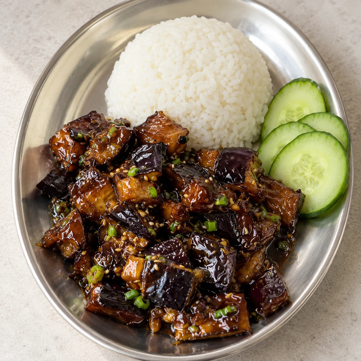 Assiette neutre avec aubergines brillantes, dôme de riz blanc cuit, quelques tranches de concombre.