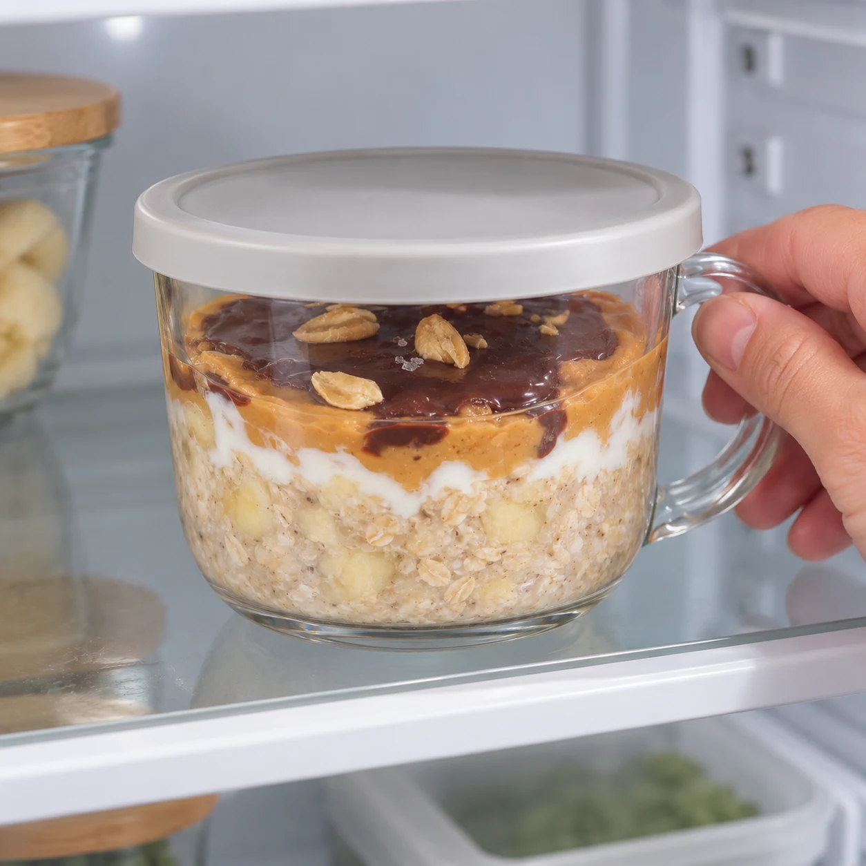 Couvrez et mettez au frais pendant 2 heures ou toute la nuit avant de déguster. Ce récipient correspond à deux petits-déjeuners ; partagez-le en deux portions. Montrer seulement cette phase de la variante à l’avoine, sans graines ni poudre, un récipient transparent à poignée sur pierre pâle, sans texte ni personne identifiable.