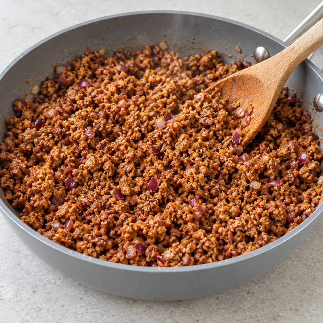 Ajoute sel, poivre, paprika, herbes de Provence et piment d’Espelette selon le goût ; mélange. Le haché cuisiné et le bouillon sont déjà salés : goûte avant d’ajouter du sel. Montrer seulement les ingrédients de cette phase ; mains anonymes, sauteuse grise et plan de travail pierre pâle.
