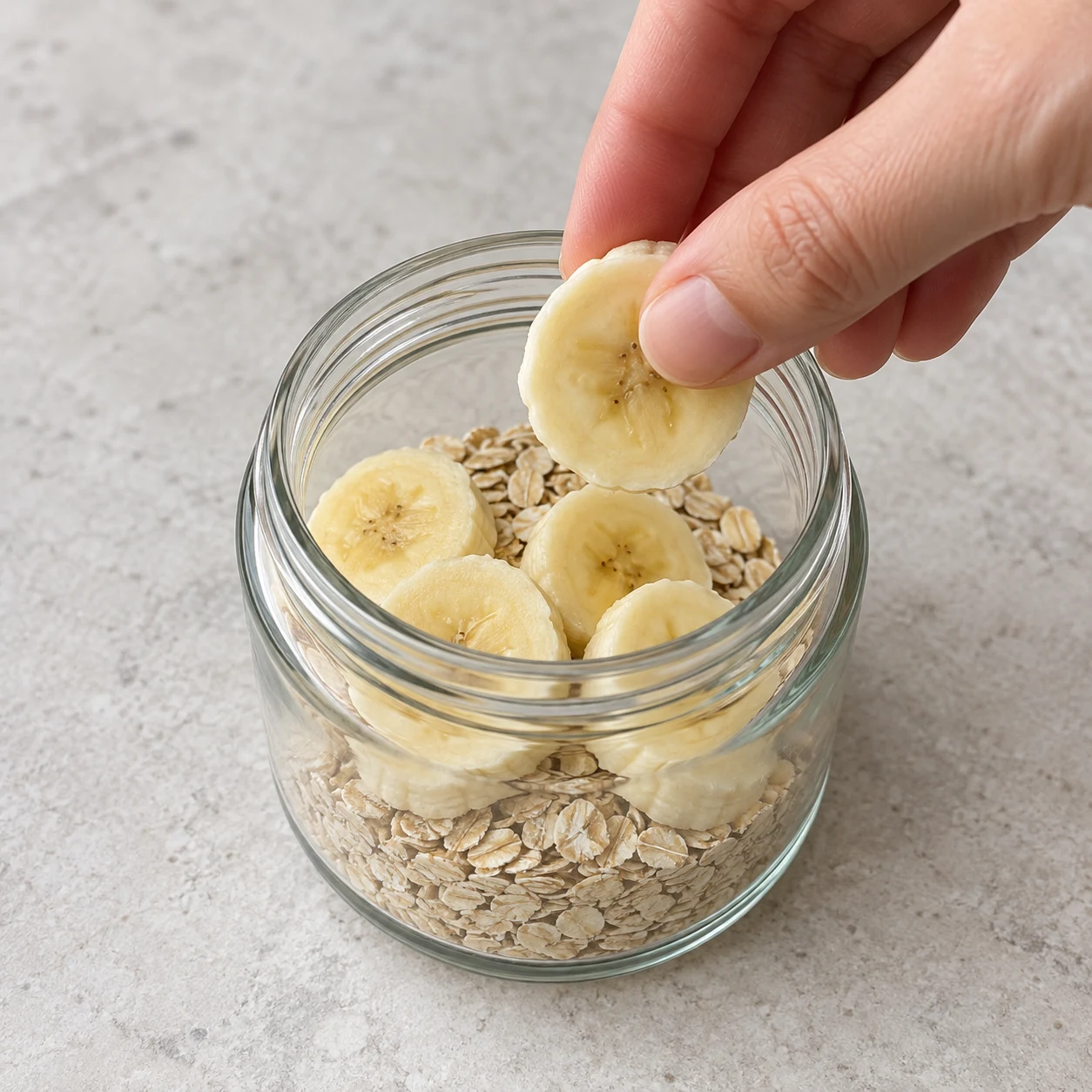 Un seul bocal contenant flocons secs et rondelles de banane, une main déposant une rondelle.