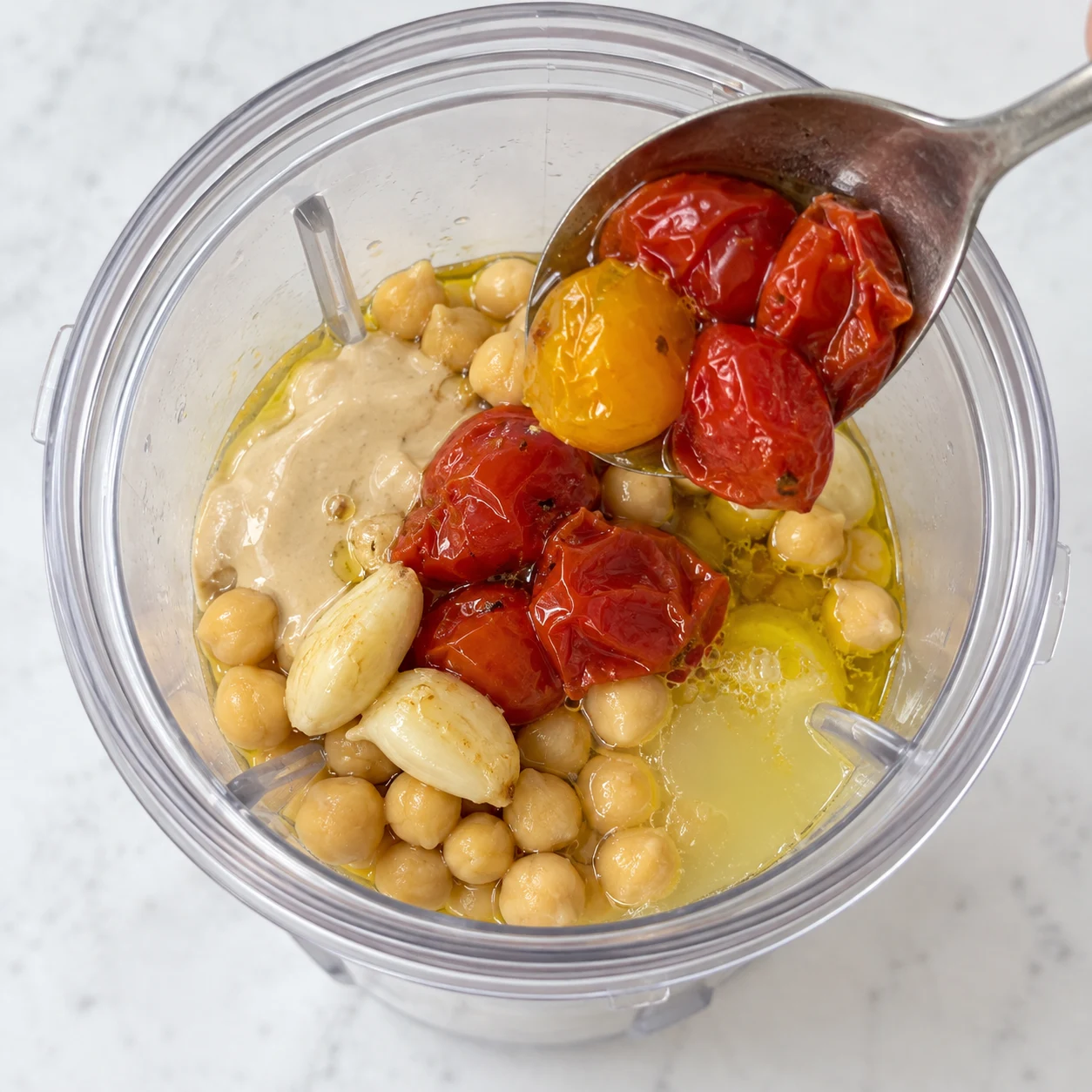 Cuillère de tomates cerises rôties entrant dans le bol du mixeur avec pois chiches, tahini, ail, citron et huile.