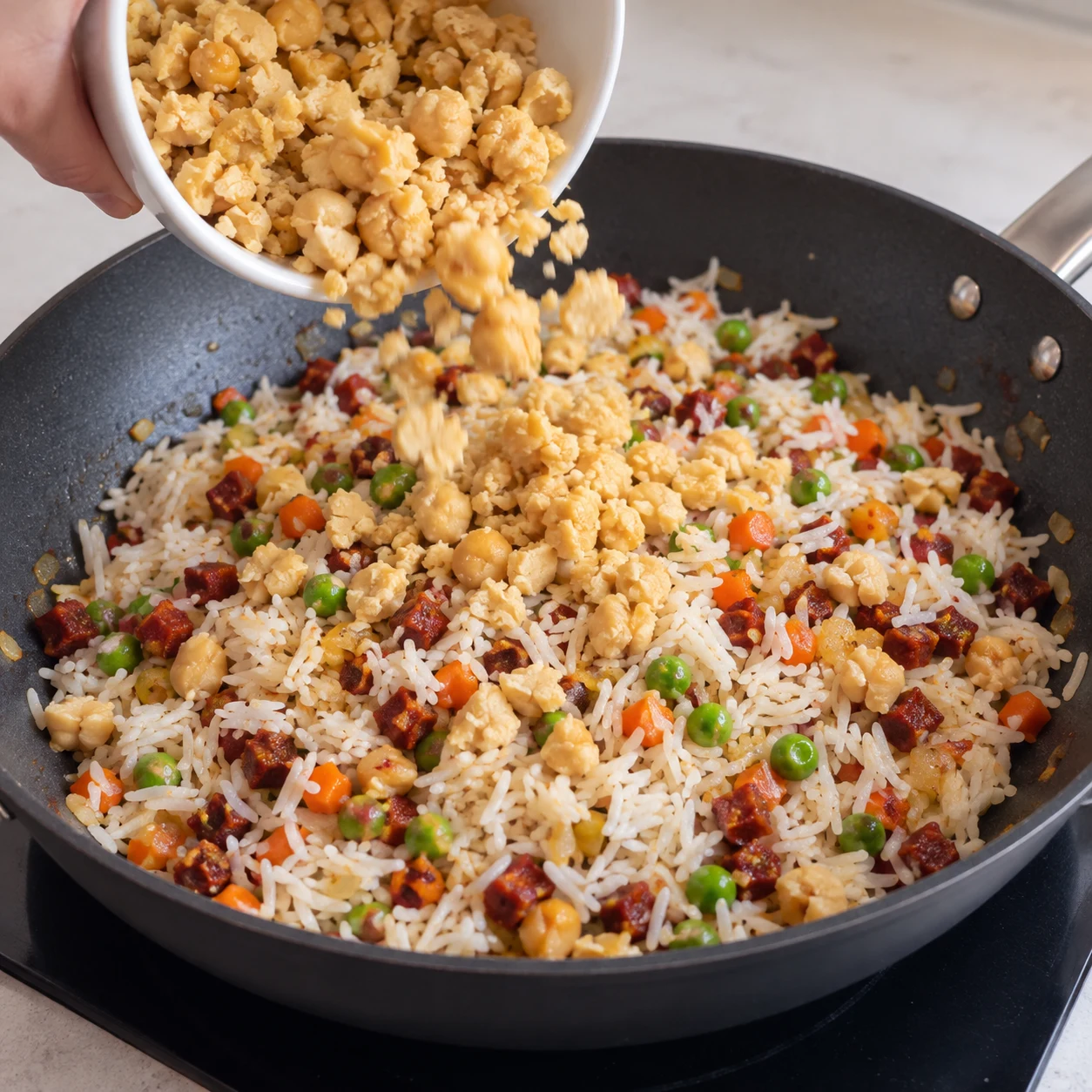 Une seule photo carrée montrant remettre les pois chiches. Les éclats de pois chiches sont répartis dans le riz. Seulement les ingrédients déjà présents : Mini chorizo végétal, Mini chorizo végétal, Huile de tournesol, Oignon jaune, Ail, Carotte, Petits pois, Riz basmati cuit et refroidi, Pois chiches cuits égouttés. Les éclats sont beige de pois chiches, sans œuf, sans tofu et sans purée lisse. Les dés sont rouge-brun de chorizo entièrement végétal, sans viande ni morceaux de gras blanc.