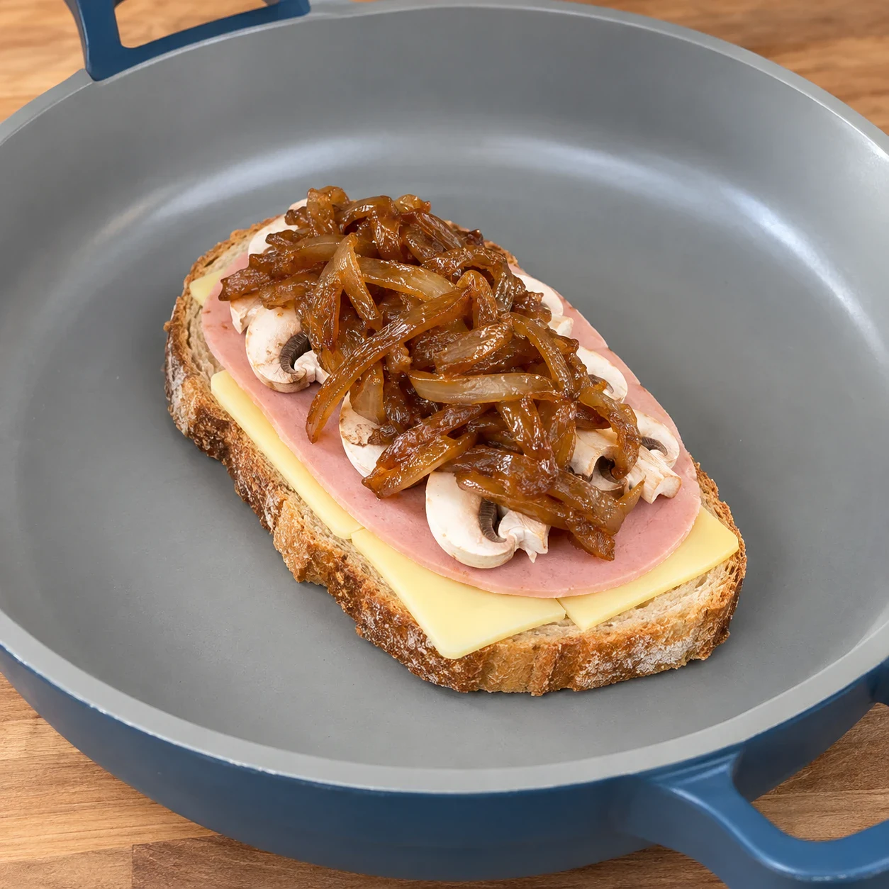 Croque ouvert dans la poêle avec tranchettes végétales jaune pâle, une tranche rose de jambon végétal, fines lamelles de champignons et oignons caramélisés.