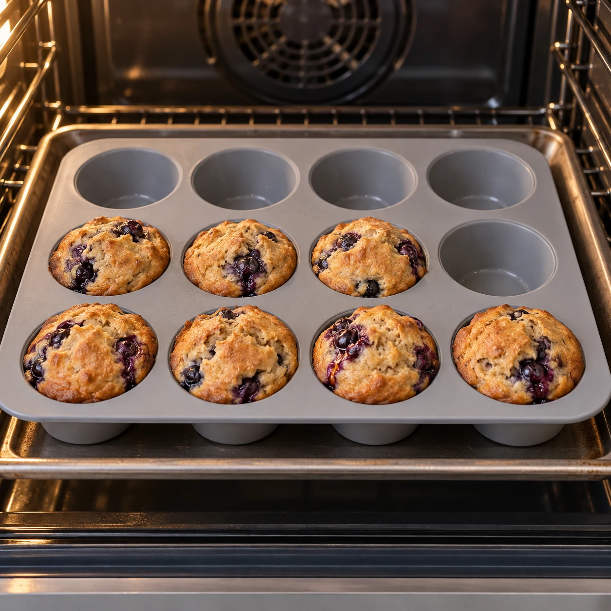 Sept muffins cuits légèrement dorés, myrtilles violettes visibles, dans un moule silicone sur une plaque.