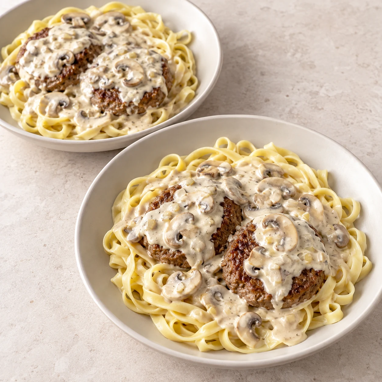 Deux assiettes contenant chacune des tagliatelles et deux steaks végétaux déjà formés, aux bords arrondis sous une sauce blanche aux champignons, sans persil ni piment encore.