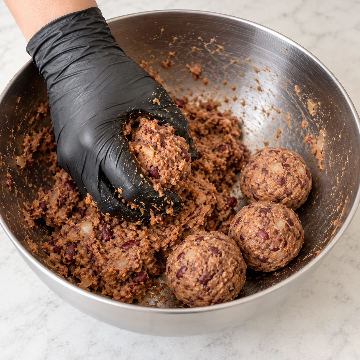 Main gantée malaxant dans un bol en inox une pâte brune de haché végétal de soja, haricots rouges écrasés, maïzena et oignon râpé.